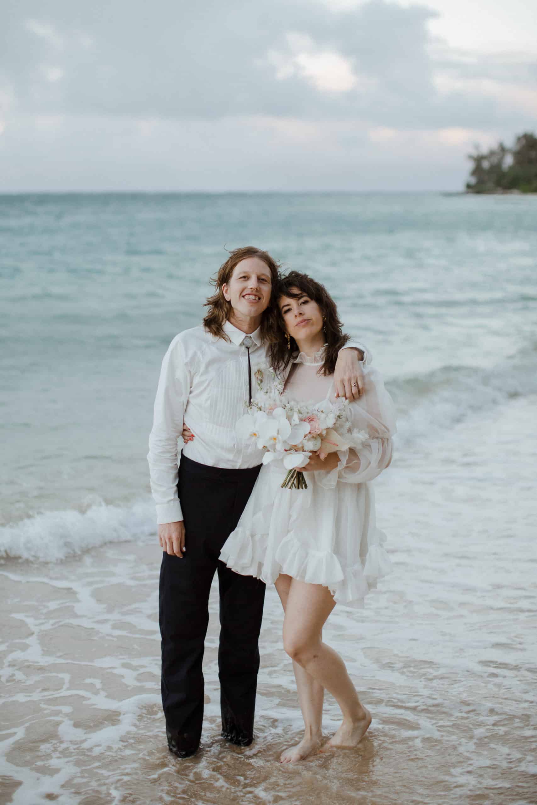 A couple stands barefoot on a beach, dressed in wedding attire. The person on the left is in a white shirt and dark pants, and the person on the right is in a white dress, holding a bouquet.