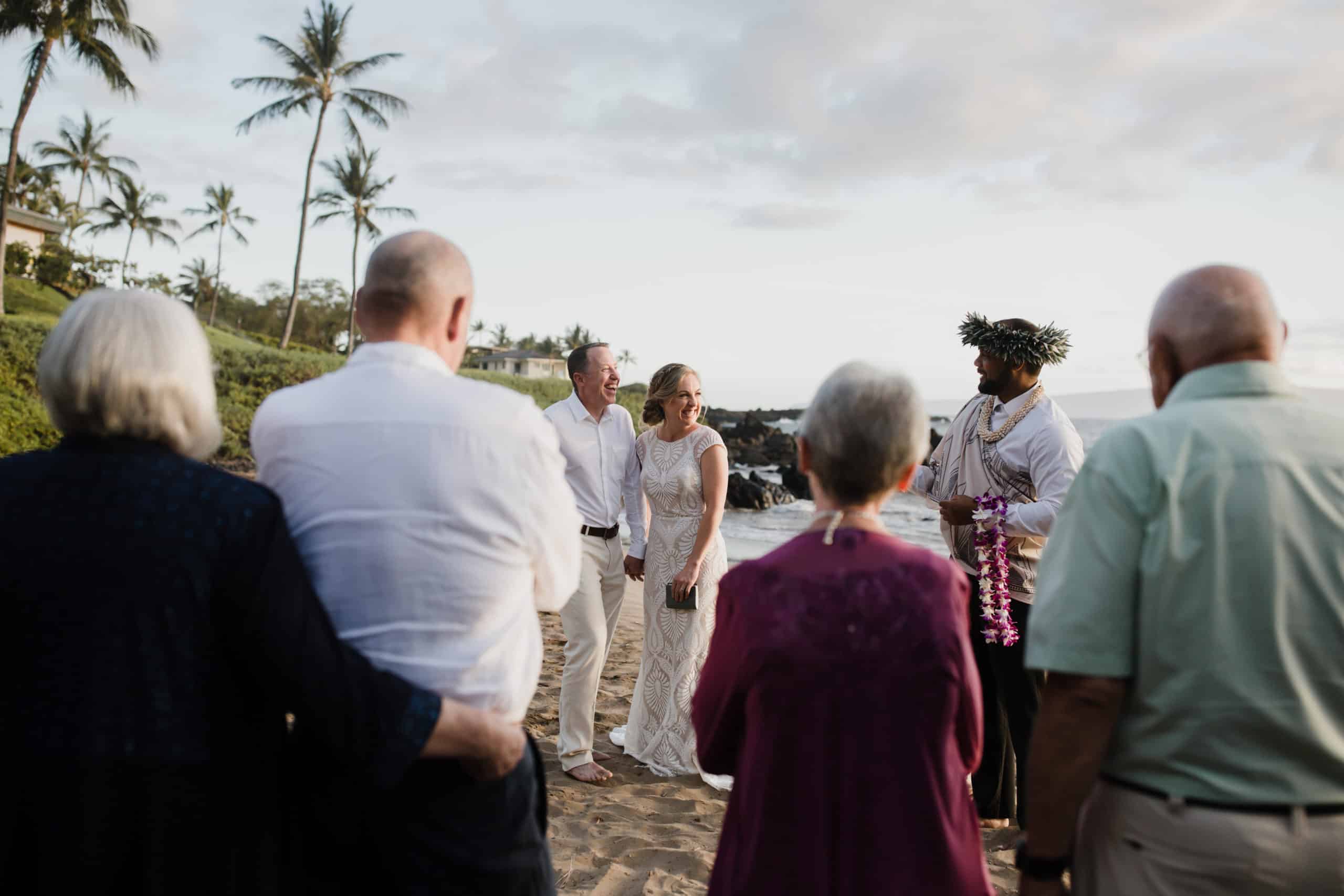 A couple stands on a beach during a wedding ceremony, surrounded by guests. A person stands in front of them holding leis. Palm trees and the ocean are visible in the background.