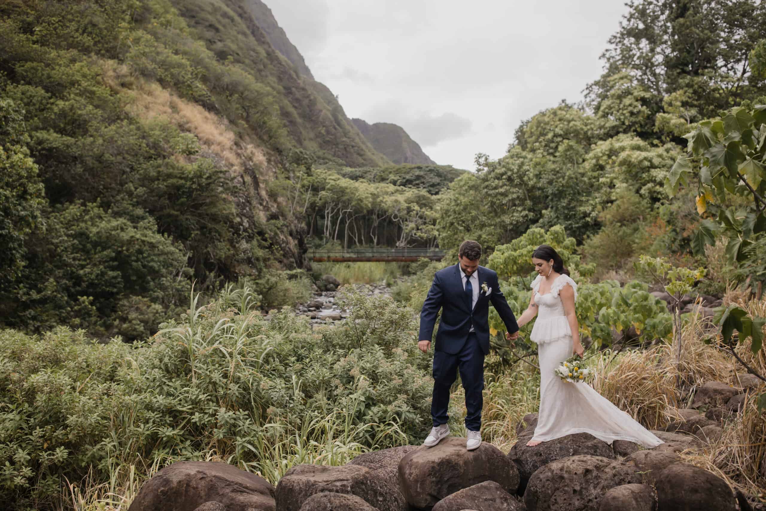 Roam Elopements, Maui Elopements, Bride and groom hold hands, walking on rocks near a stream. Intimate Hawaii wedding.