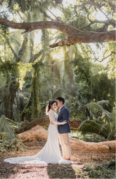 A couple stands in a forest, embracing under a large tree branch. The bride wears a white gown, and the groom is in a suit. Sunlight filters through the trees.