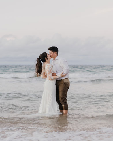 A couple kisses while standing in the shallow ocean water, with the beach and cloudy sky in the background.