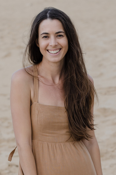 A woman with long hair smiles while standing on a sandy beach, wearing a sleeveless tan dress.