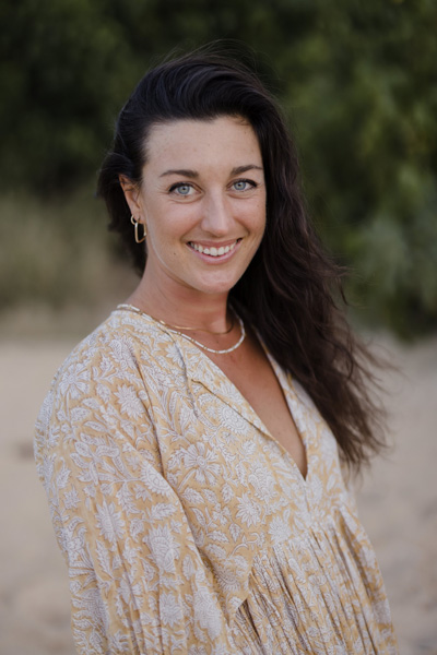 A person with long dark hair and a floral-patterned beige top stands outdoors, smiling at the camera.