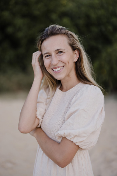 A person in a light-colored dress stands outdoors, smiling, with one hand touching their hair. Trees and sand are in the background.