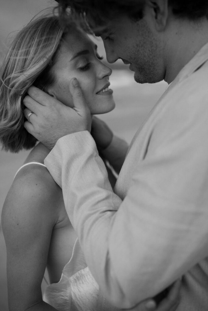 A couple embracing closely, with the man's hands gently holding the woman's face, both looking at each other affectionately. The image is in black and white.