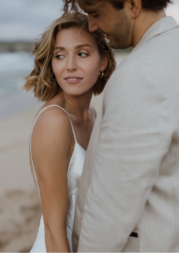 A couple stands close together on a beach. The woman in a white dress looks over her shoulder, while the man in a light-colored suit faces her.