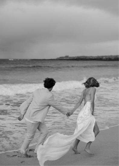 A couple in formal attire holds hands and runs along a beach shoreline with waves in the background.