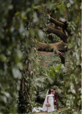 A couple in wedding attire stands close together, surrounded by lush greenery and large trees.