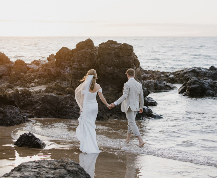 A couple walks hand in hand along a rocky beach, with the ocean and sunset in the background.