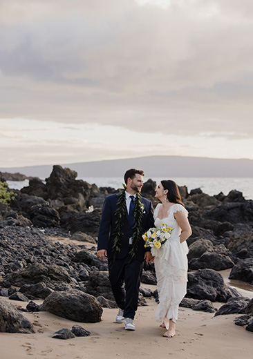 A bride and groom walk hand in hand on a rocky beach, with the ocean in the background. The bride holds a bouquet of yellow and white flowers.