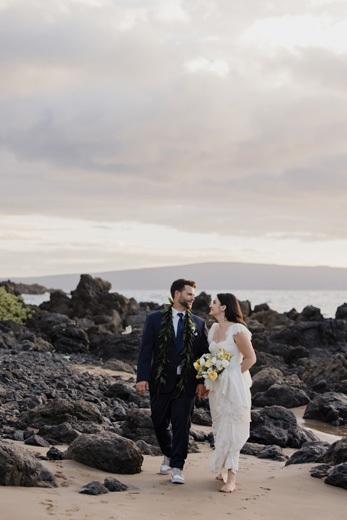 A couple in wedding attire walks barefoot on a rocky beach, holding hands and smiling at each other. The bride carries a bouquet of yellow and white flowers. Cloudy sky in the background.