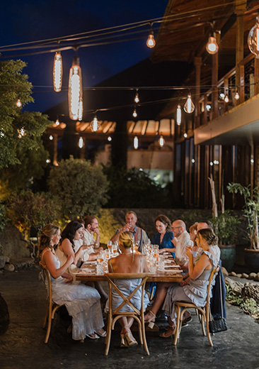 A group of people sit around a dinner table outdoors, surrounded by warm lights and greenery, during the evening.