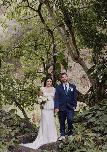 A bride and groom stand outdoors in a lush, green forest setting. The bride holds a bouquet of flowers.