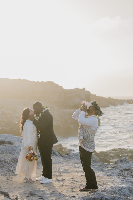 A couple kisses during a beach wedding ceremony as an officiant, wearing a floral crown, blows into a conch shell. The ocean and rocky shore are in the background.