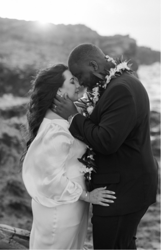 A couple embraces forehead-to-forehead, wearing floral garlands by the ocean. The setting is romantic with a serene backdrop.