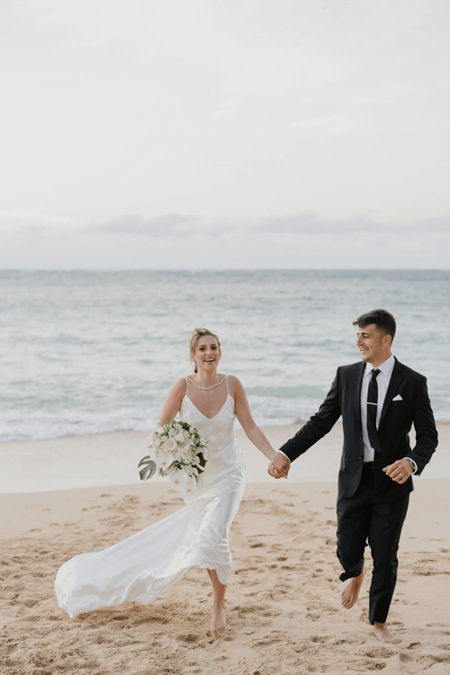 A bride in a white dress and a groom in a suit holding hands, running barefoot on a sandy beach with the ocean in the background.