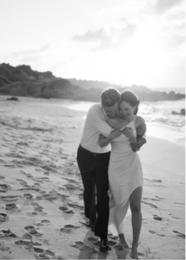 A couple walks closely together on a sandy beach at sunset, leaving footprints behind.