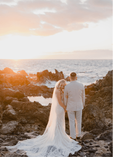 A bride and groom stand on rocky terrain by the ocean at sunset, holding hands. The bride wears a lace gown with a long veil, and the groom is in a light-colored suit.