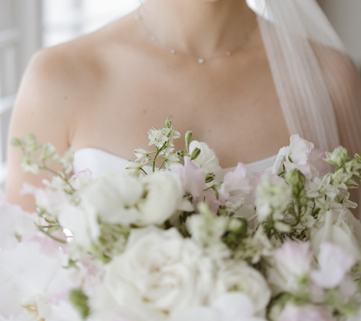 A bride in a white dress holding a bouquet of white and light pink flowers, with a veil draped over her shoulders.