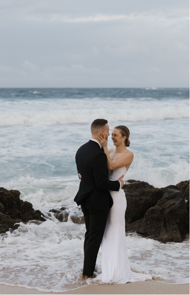 A couple in formal attire embraces on a rocky beach with ocean waves in the background.