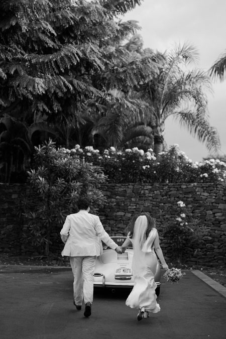 A couple in wedding attire walks hand in hand toward a vintage car with "Just Married" on the rear, surrounded by lush greenery.