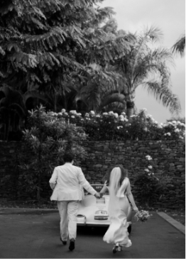 A couple in formal attire walks hand in hand towards a classic car. The scene is set outdoors, surrounded by lush greenery and flowers.