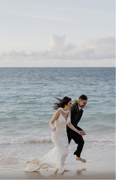 A bride in a white dress and a groom in a suit joyfully run barefoot along the beach near the ocean.