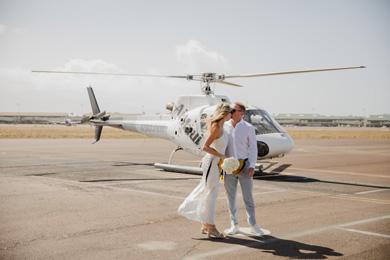 A couple stands together on an airport tarmac with a helicopter in the background. The woman holds a bouquet.