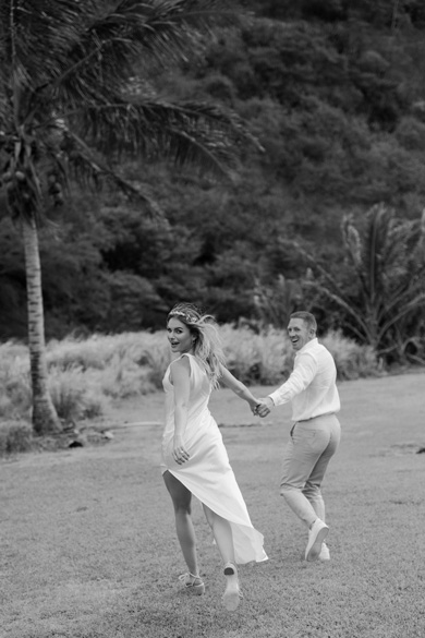 A couple holding hands, running joyfully across a grassy field with palm trees and greenery in the background. The woman wears a flowing white dress, and the man wears a light-colored outfit.