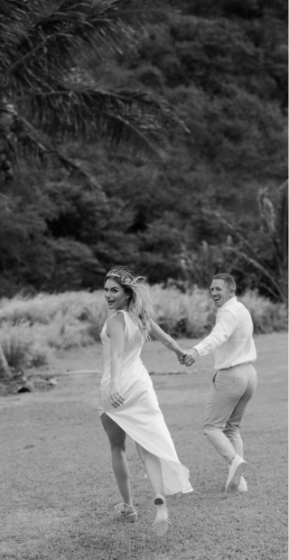 A couple in formal attire holds hands and walks across a grassy field, smiling and looking back.