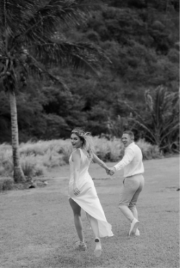 A couple in wedding attire holding hands and running outdoors, with trees and grass in the background. The woman looks back while smiling. Black and white image.