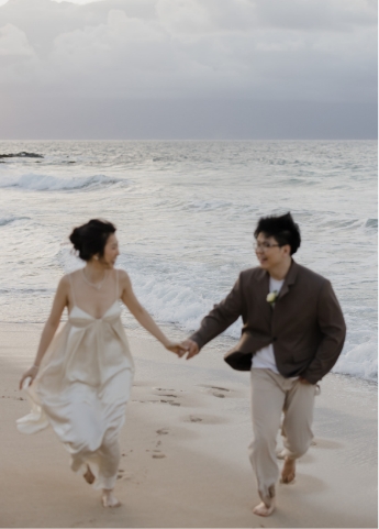 A couple runs barefoot on a beach, holding hands, with the ocean waves in the background and an overcast sky.