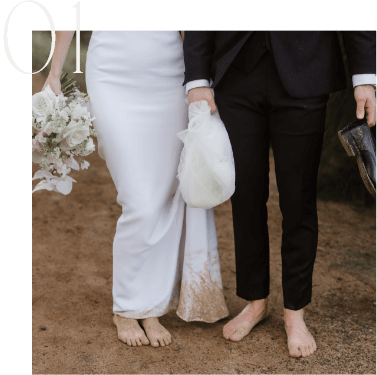 A couple in wedding attire stand barefoot on a dirt path. The woman holds a bouquet of flowers and her dress train. The man carries his shoes.