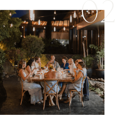 A group of people sit around a round wooden table, dining outdoors in an evening setting with ambient lighting.