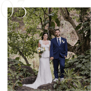 Bride and groom in wedding attire stand together on a rocky path, surrounded by lush greenery.