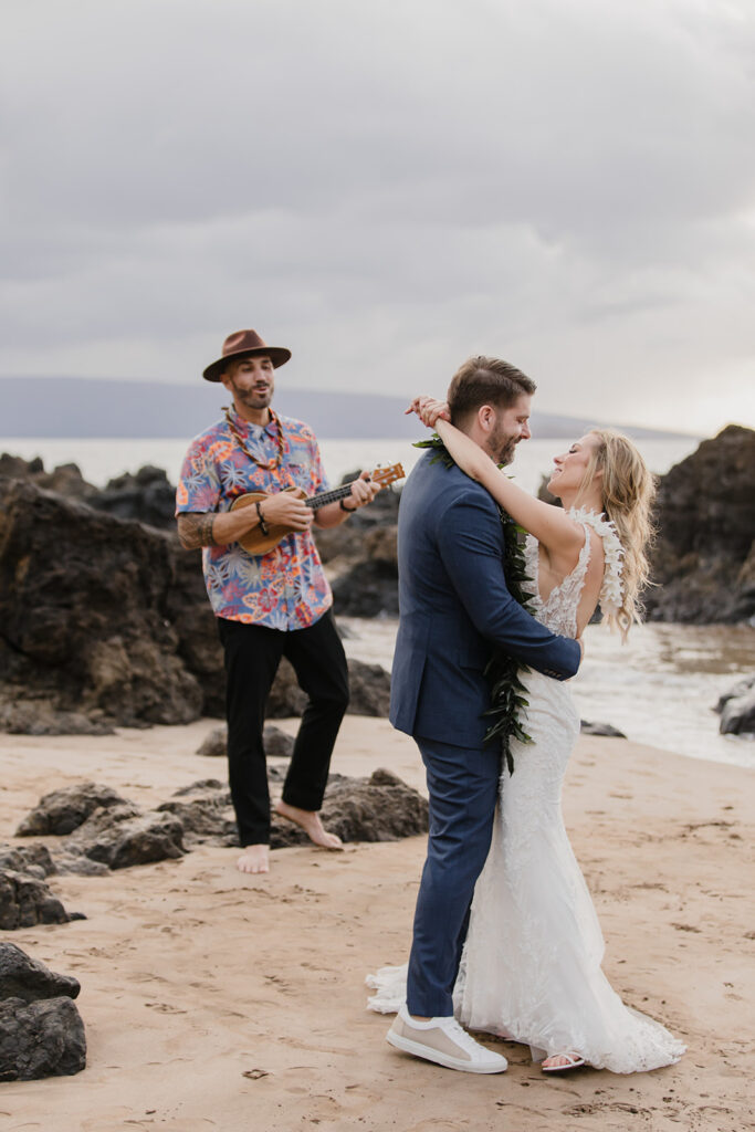 Roam Elopements, Maui Elopements, Bride and groom embrace on beach, musician plays ukulele