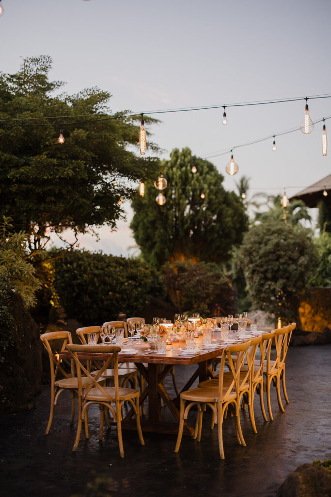 Roam Elopements, Maui Elopements, Elegant outdoor dinner party with string lights, wooden cross-back chairs, and a long wooden table set for many guests.