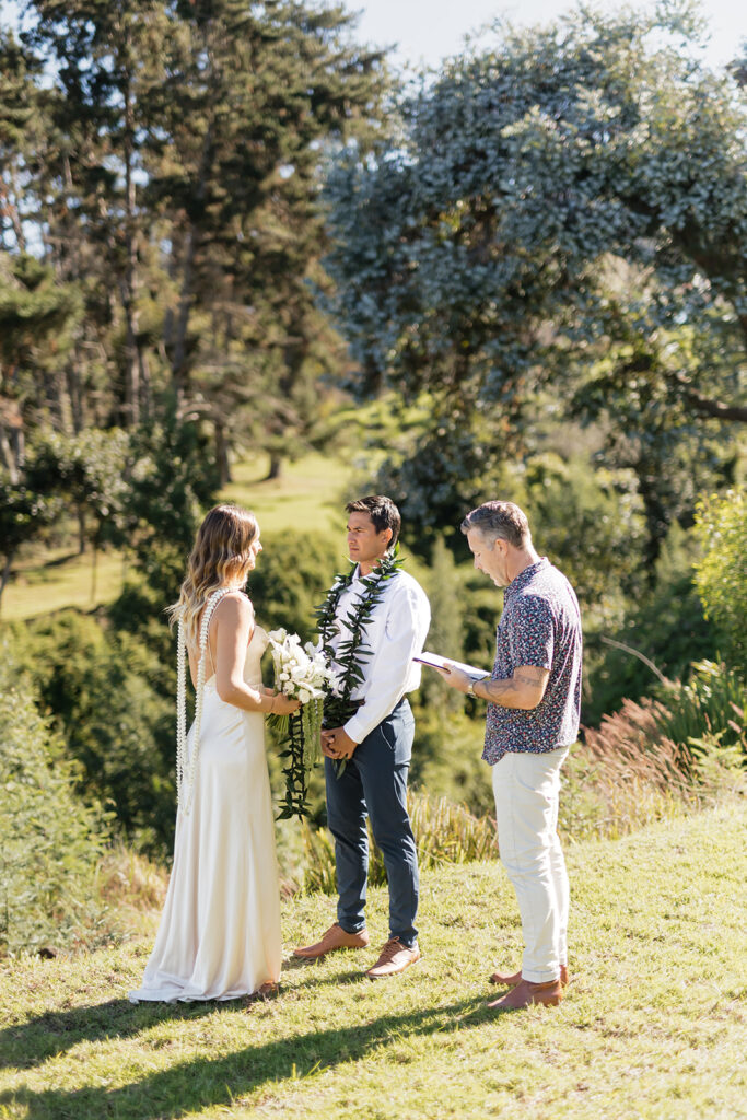 Roam Elopements, Maui Elopements, Elopement ceremony: Bride in white dress, groom in lei, officiant with notes in a lush outdoor setting.