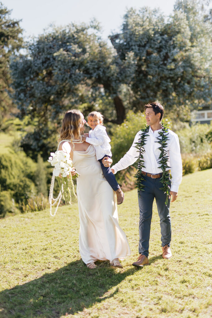 Roam Elopements, Maui Elopements, Family elopement photo: Bride, groom with lei, and child on lawn.