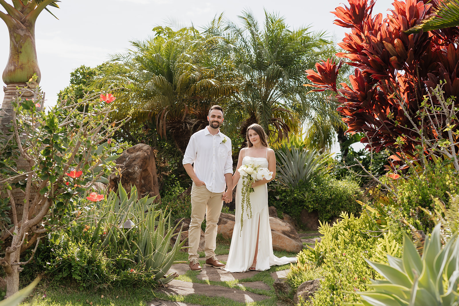 Roam Elopements, Maui Elopements, Eloping couple in tropical garden setting. Bride holding bouquet, groom in linen shirt.