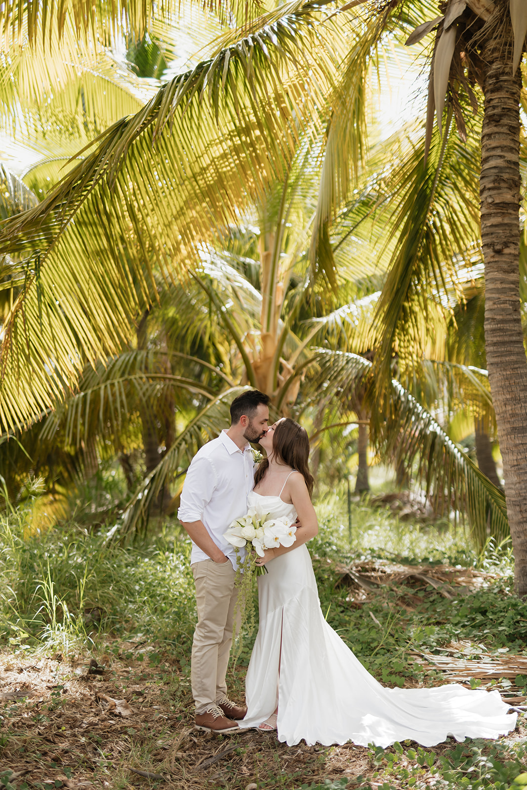 Roam Elopements, Maui Elopements, Romantic elopement: Couple kisses under palm trees. Bride in white dress holds lilies.