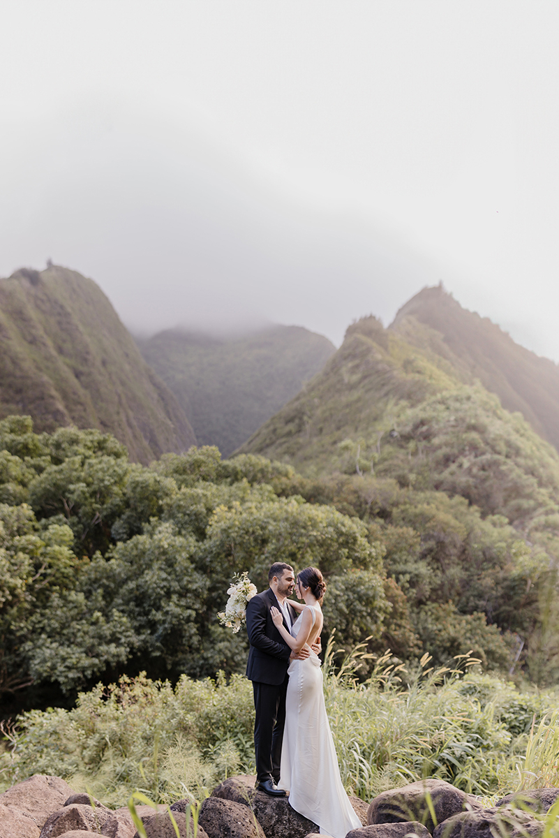 Argine-Artur-Maui-Elopement-24-cropped - Roam Elopements Roam Elopements, Maui Elopements, Bride and groom embrace on a rocky outcrop overlooking lush green mountains.