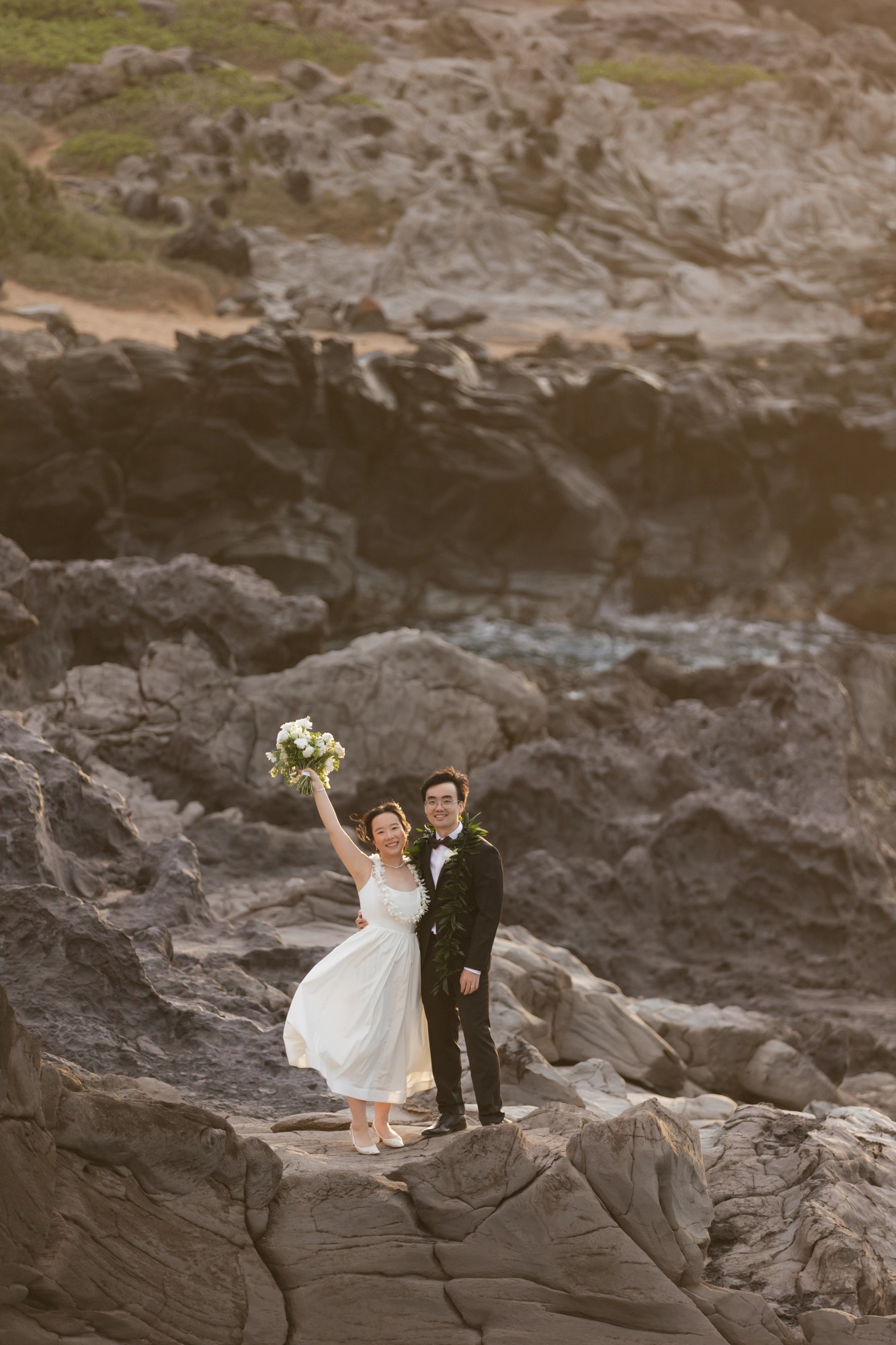 Zhenyi Hanqi - Roam Elopements Roam Elopements, Maui Elopements, Happy newlyweds pose on rocky coastline, bride holding bouquet.