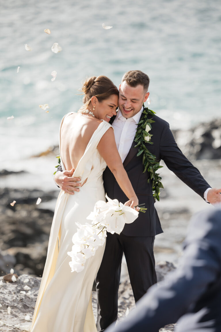 A bride and groom hug by the shore as petals drift around them, ocean in the background.