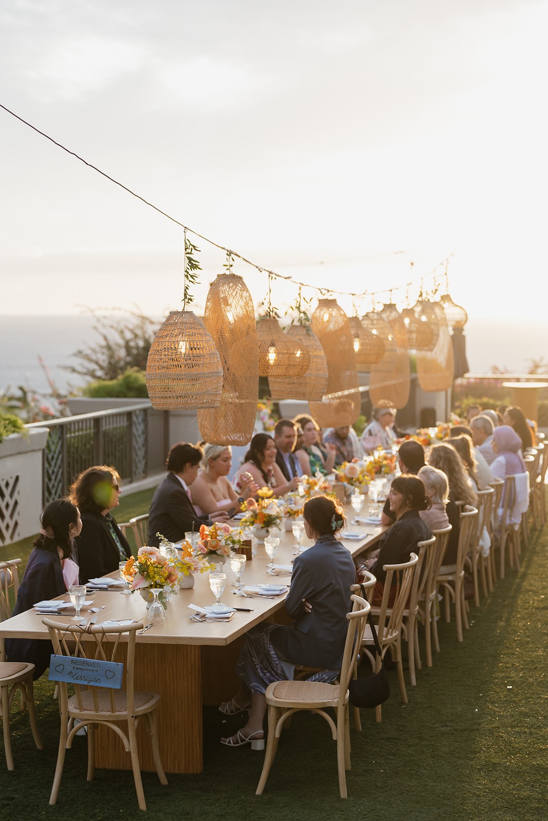 Outdoor long dinner table at sunset, guests seated with floral centerpieces and wicker lanterns overhead.",
