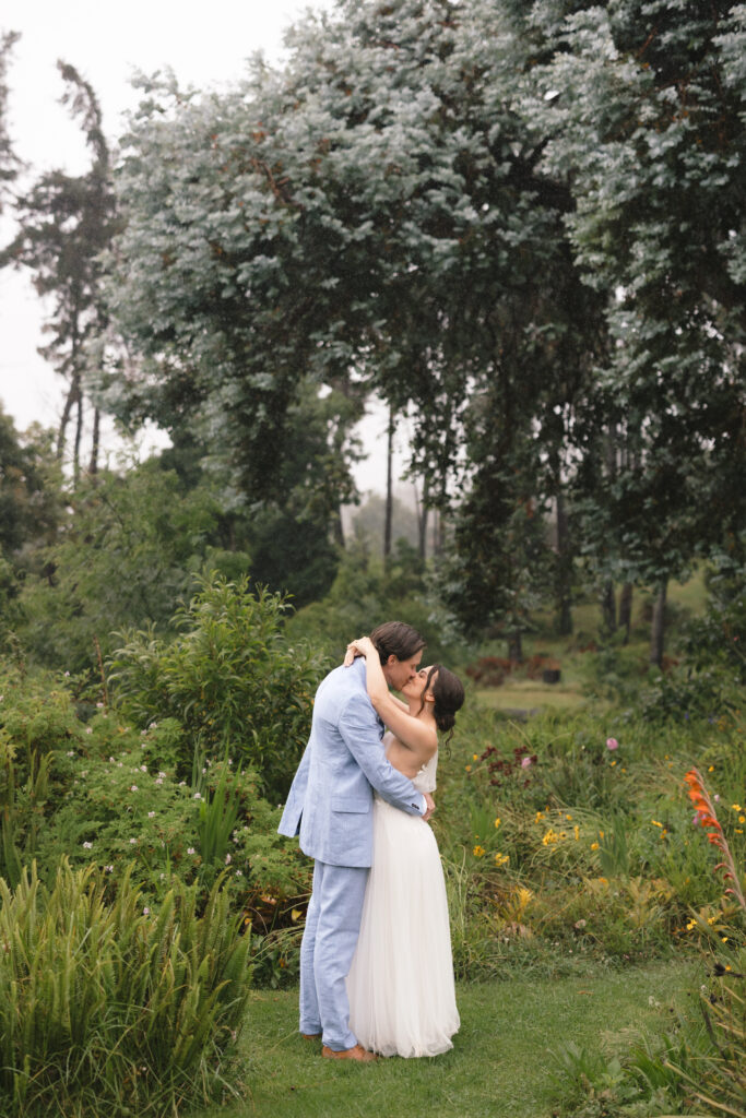 Isa Matt - Roam Elopements A couple in wedding attire embracing in a garden with greenery and tall trees in the background.