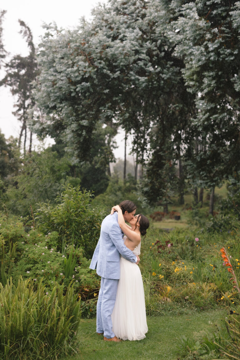 A couple in wedding attire embracing in a garden with greenery and tall trees in the background.